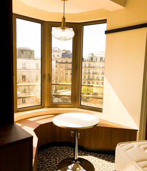 Vue de la fenêtre sur les immeubles haussmaniens de Paris, il y a une assise confortable et une petite table ainsi qu'un lustre en verre