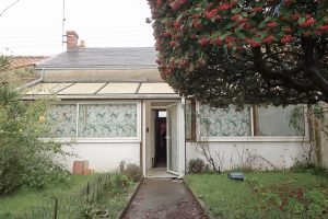 Maison avant travaux avec allée en béton traversant le jardin remplit de verdure. Il y a un arbre et une vue sur la véranda de la maison avec des cheminées.