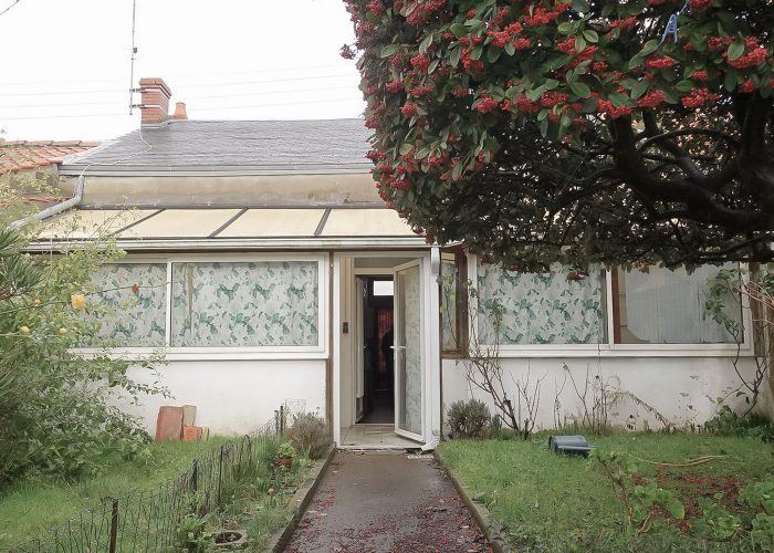 Maison avant travaux avec allée en béton traversant le jardin remplit de verdure. Il y a un arbre et une vue sur la véranda de la maison avec des cheminées.